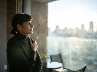 Woman with dark hair and a black turtleneck sweater standing near a large window, looking thoughtfully at a sunny city skyline view in...