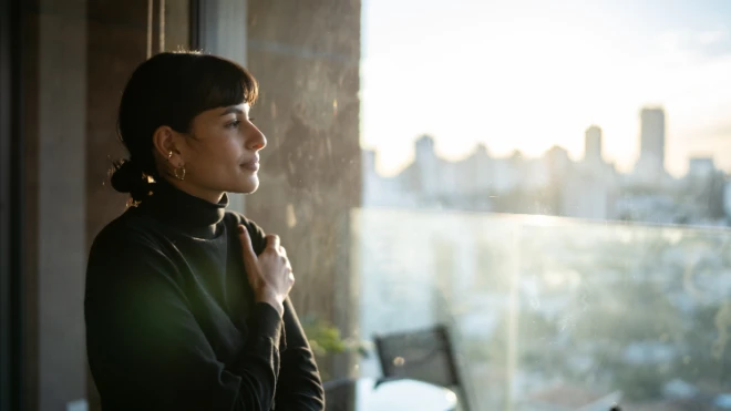 Woman with dark hair and a black turtleneck sweater standing near a large window, looking thoughtfully at a sunny city skyline view in...