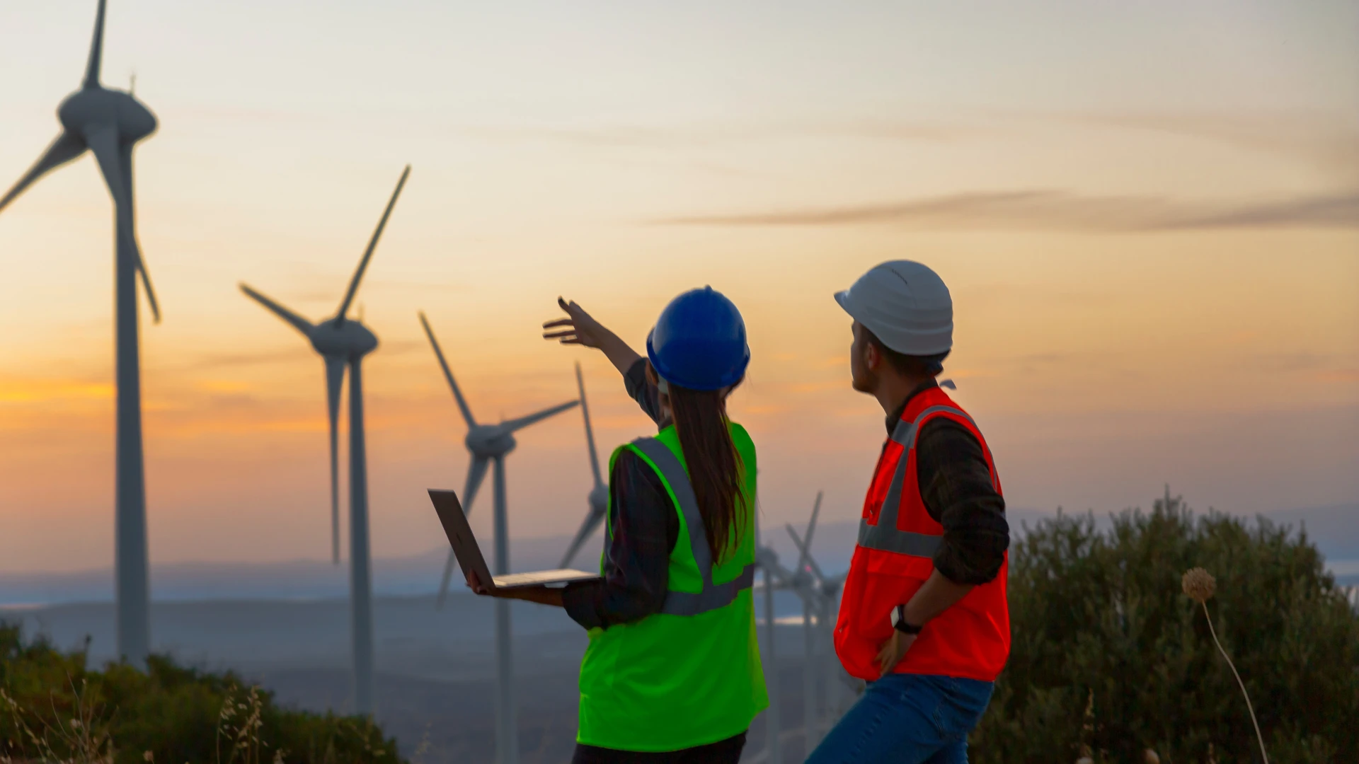 Two wind energy technicians (green vest/blue hard hat pointing with laptop; orange vest/white hard hat) overlook a field of...