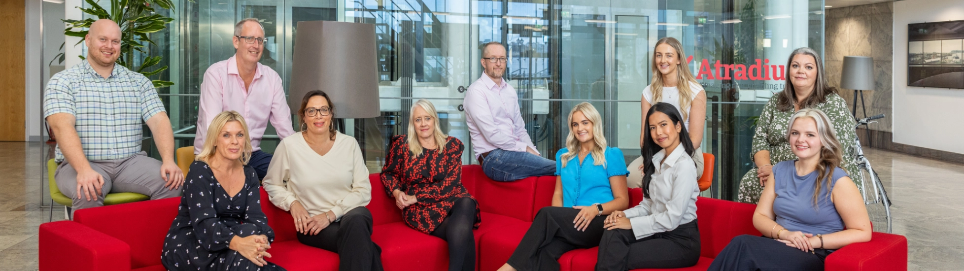 A group shot of the Atradius CST team, they are sat on a red sofa together in the atrium of the Cardiff office